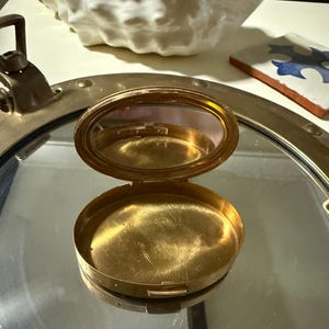 May include: A small, oval-shaped gold-toned metal compact case with a hinged lid. The interior features a polished gold base and a mirror attached to the inside of the lid. The compact is open, revealing its reflective surfaces and metallic finish.