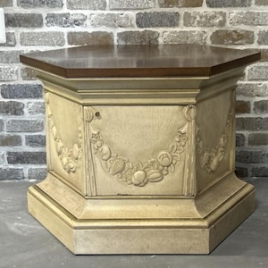May include: A hexagonal side table with a dark brown top and a cream-colored base. The base features carved floral and fruit garland designs. The table is approximately 24 inches tall.