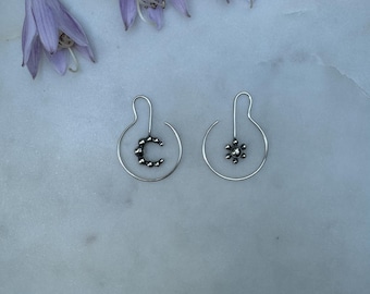 May include: A pair of silver earrings, one with a crescent moon design and the other with a sunburst design. The earrings are displayed on a white marble surface, with several purple flowers in the background.