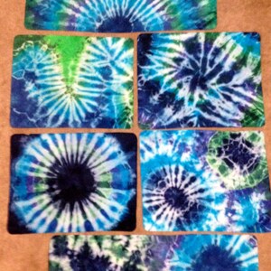 May include: Five square tie-dye fabric coasters in shades of blue, green, and white. The coasters feature a circular pattern with a dark blue center.
