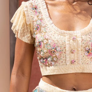 May include: A cream-colored crop top with a lace design and floral embroidery. The top has a scoop neckline and short, ruffled sleeves. The embroidery features pearls, sequins, and beads in shades of pink, green, and blue.