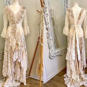 May include: Ivory-colored, long-sleeved dress with a V-neck and layered, ruffled skirt. The dress features lace and ribbon details, and a matching sash. The dress is displayed on a mannequin, with a mirror and decorative stand in the background.