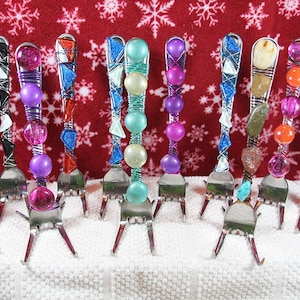 May include: A collection of 12 decorative forks with colourful beads and wire wrapped around the handle. The forks are arranged in a row on a white surface.