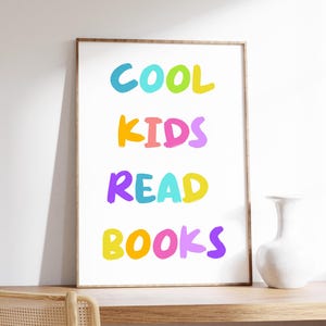 May include: A framed print with the words "COOL KIDS READ BOOKS" in bright, colorful letters. The text is in a playful font, with each word in a different color. The frame is a light brown color, and the print is displayed on a wooden surface.