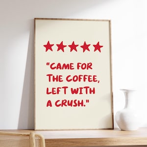 Op de afbeelding: Een ingelijste print met een crèmekleurige achtergrond en rode tekst met de tekst "CAME FOR THE COFFEE, LEFT WITH A CRUSH." Boven de tekst staan vijf rode sterren. De lijst is lichtbruin.