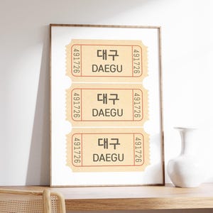 May include: A framed print featuring three vintage-style tickets. Each ticket is tan with red borders and the text "DAEGU" in Korean and English. The tickets have the number "491726" printed on each side. The frame is brown.