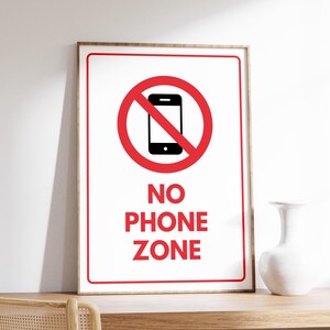 Puede incluir: Un cartel blanco con un círculo rojo y una línea roja diagonal que atraviesa una silueta negra de un teléfono inteligente. El texto "NO PHONE ZONE" está escrito en letras rojas debajo del círculo.