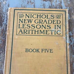 May include: A vintage book titled "Nichols New Graded Lessons in Arithmetic Book Five" by Thompson Brown Co. The book has a tan cover with green text and a green border.