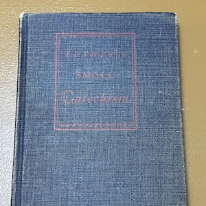May include: A vintage book titled "Luther's Small Catechism" with a blue cover. The title is in red lettering within a red rectangular frame. The book's cover has a textured appearance.