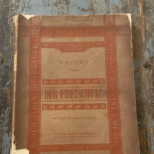 May include: A vintage book cover for the opera "Der Freischütz" by Carl Maria von Weber. The cover is brown with red accents and features the title in large, bold letters. The book is part of the Novellos Original Octavo Edition and was edited by Natalia Macfarren. The cover also includes the names of other composers, including Mozart, Beethoven, Spohr, Rossini, and Auber.