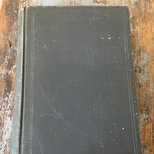 May include: A dark gray hardcover book with a textured cover. The book has a rectangular shape and is lying on a wooden surface. The edges of the book are slightly worn, suggesting age or use.