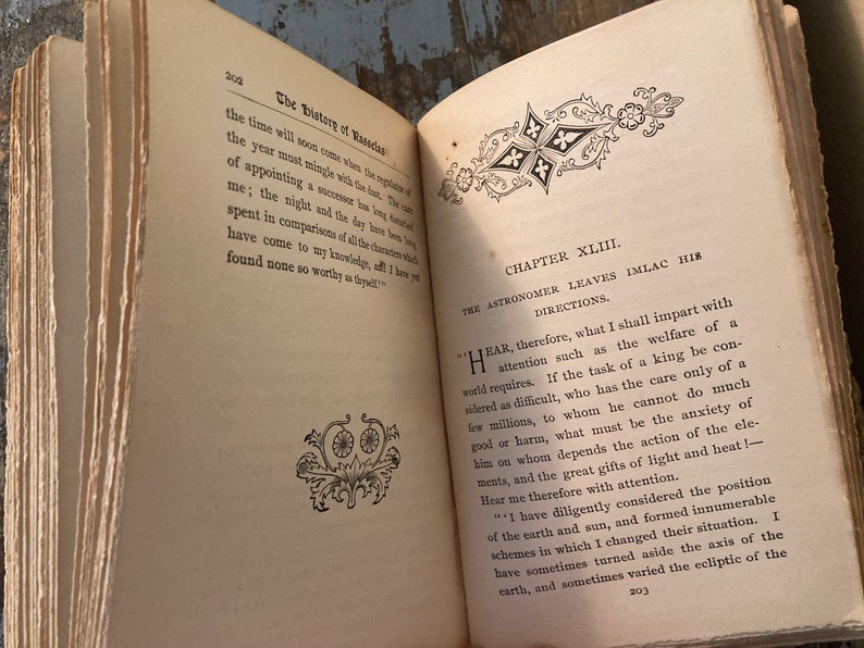 May include: An open book with yellowed pages and a decorative design on the cover. The book is titled "The History of Rasselas" and the chapter is titled "The Astronomer Leaves Imlac His Directions". The text on the page discusses the responsibilities of a ruler and the importance of considering the welfare of the people.