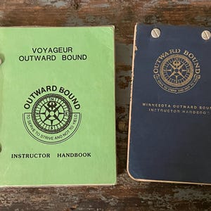 May include: Two green and blue instructor handbooks for Outward Bound, a youth development organization. The front cover of the green book says "Voyageur Outward Bound" and features a compass with the words "To serve to strive and not to yield". The front cover of the blue book says "Minnesota Outward Bound Instructor Handbook".