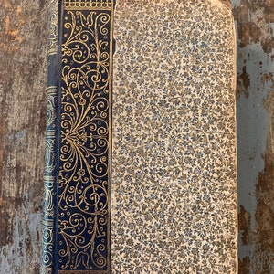 May include: A vintage book with a dark blue cover and gold-colored floral designs. The book has a patterned paper cover with a floral design.