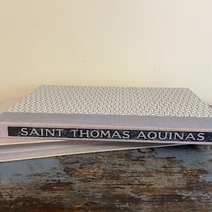 May include: A hardback book with a white and black patterned cover and a gray spine. The title "SAINT THOMAS AQUINAS" is printed in black on the spine.