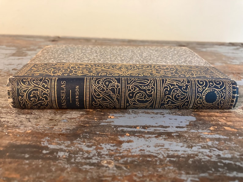 May include: A vintage hardcover book with a dark blue cover and gold lettering. The title "Rasselas" and the author "Johnson" are visible on the spine. The book has an intricate gold design on the cover.