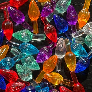 May include: Assortment of colorful plastic Christmas light bulb covers in various shades of red, orange, yellow, green, blue, purple, and clear. The bulbs are arranged in a random pattern on a black surface.