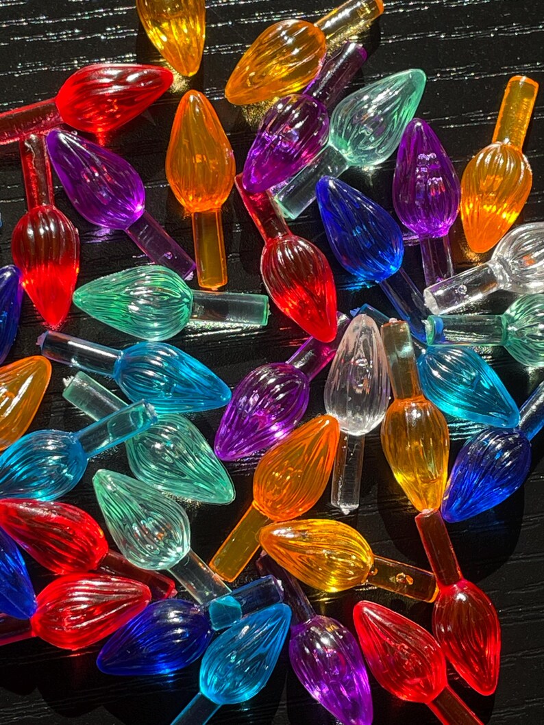 May include: Assortment of colorful plastic Christmas light bulb covers in various shades of red, orange, yellow, green, blue, purple, and clear. The bulbs are arranged in a random pattern on a black surface.