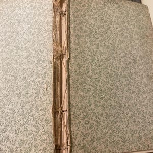 May include: Close-up of the spine of a vintage book with a floral pattern on the cover. The book is bound with a brown thread and has a worn, aged appearance.