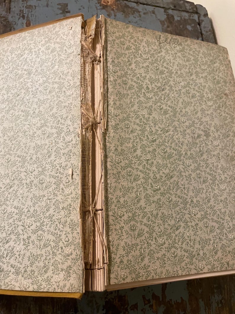 May include: Close-up of the spine of a vintage book with a floral pattern on the cover. The book is bound with a brown thread and has a worn, aged appearance.