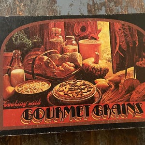 May include: A vintage cookbook with a red cover and the title "Cooking with Gourmet Grains". The cover features a still life of bread, fruit, and a pie.