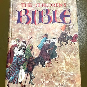 May include: A vintage hardcover book titled "The Children's Bible." The cover features an illustration of people in robes riding donkeys, with the title in large, stylized letters. The background has a textured, aged appearance.