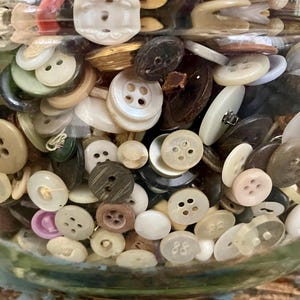 May include: A glass jar filled with a variety of buttons in different colors, shapes, and sizes. The buttons are mostly white, brown, and black, with some red, pink, and green buttons.