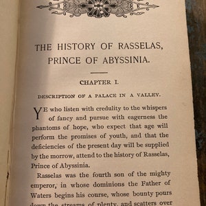 May include: A vintage book with a decorative border and the title "The History of Rasselas, Prince of Abyssinia." The book is open to the first chapter, "Description of a Palace in a Valley."