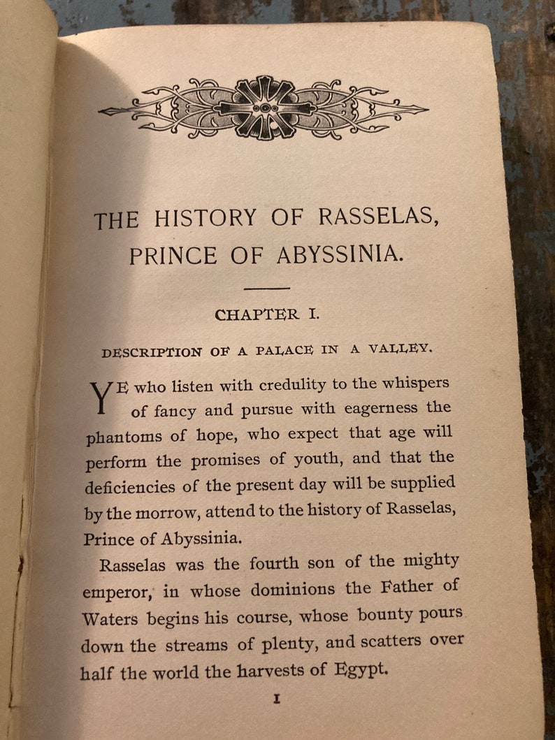 May include: A vintage book with a decorative border and the title "The History of Rasselas, Prince of Abyssinia." The book is open to the first chapter, "Description of a Palace in a Valley."