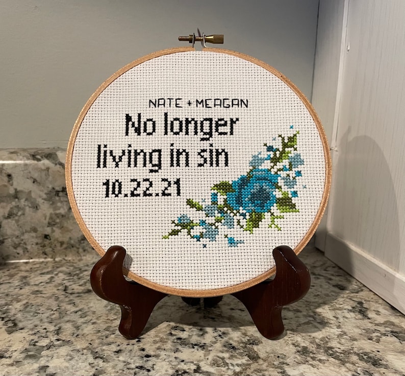 Puede incluir: Un aro de bordado blanco con un dise&ntilde;o floral azul y verde. El aro tiene el texto "Nate + Meagan No longer living in sin 10.22.21" bordado en &eacute;l.