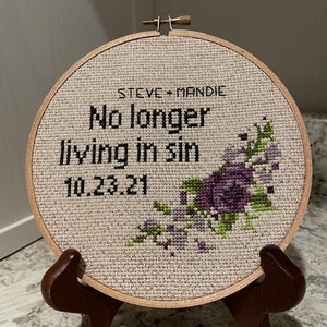 Puede incluir: Un aro de bordado redondo de madera con un fondo de tela beige. La tela tiene un dise&ntilde;o de punto de cruz de flores moradas y hojas verdes. El texto "STEVE + MANDIE No longer living in sin 10.23.21" est&aacute; bordado en hilo negro.