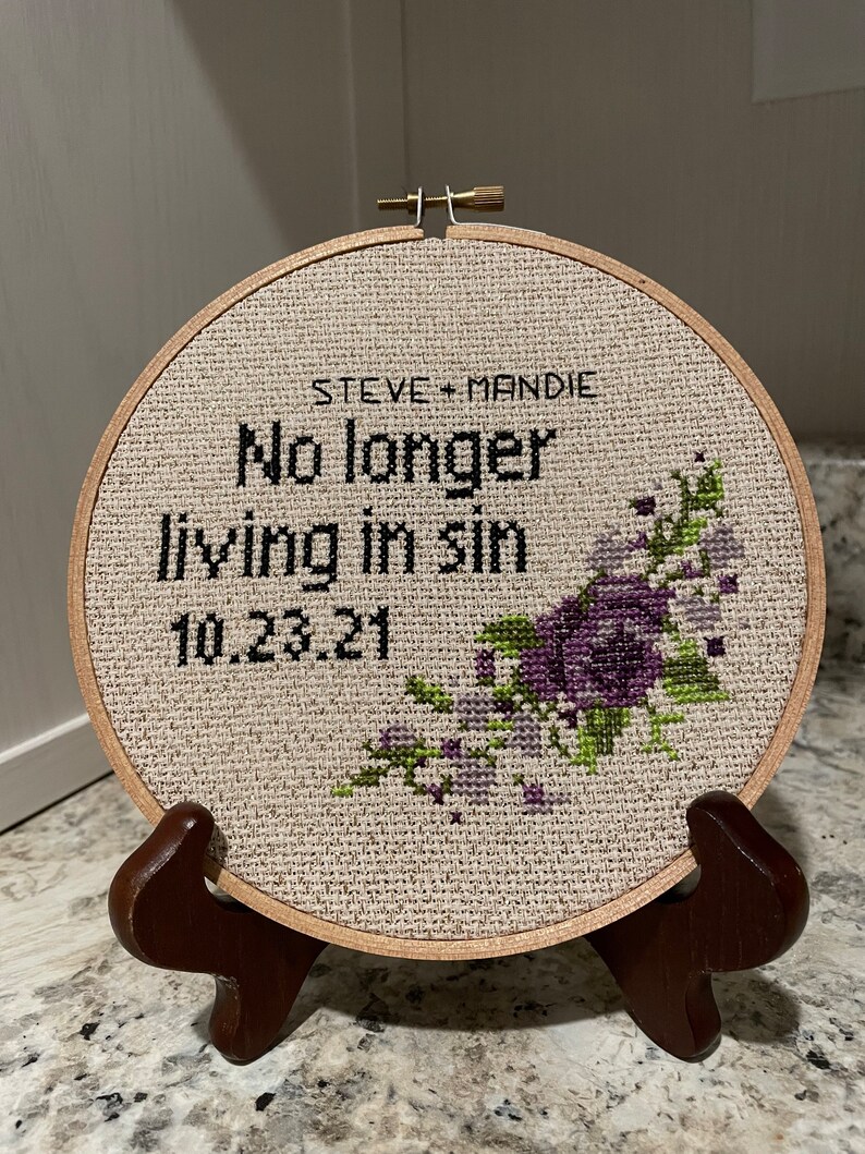 Puede incluir: Un aro de bordado redondo de madera con un fondo de tela beige. La tela tiene un dise&ntilde;o de punto de cruz de flores moradas y hojas verdes. El texto "STEVE + MANDIE No longer living in sin 10.23.21" est&aacute; bordado en hilo negro.