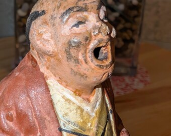 May include: A weathered, antique figurine of a man with an open mouth, possibly singing or shouting. The figure wears a reddish-brown robe over a yellow garment. The face has visible details, including dark eyebrows and a textured surface. The figurine is likely ceramic or a similar material.