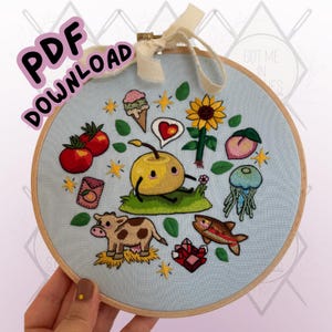 Puede incluir: Un bastidor de bordado azul claro con un diseño caprichoso bordado. El diseño incluye un personaje de fruta amarilla sonriente, una vaca, tomates, un girasol y el texto "PDF DOWNLOAD".