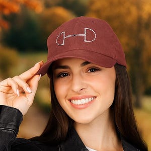 May include: A maroon baseball cap with a silver horse bit design. The cap is made of corduroy material and has a curved brim. The hat is being held up by a person.