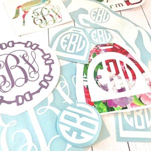 May include: A collection of monogram decals in various shapes and sizes. Designs include a floral horse, a circular monogram, and monograms within a saddle shape. Colors include white, light blue, purple, and floral patterns with red, pink, and green.