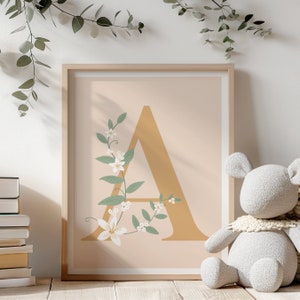 Może przedstawiać: Wydruk w ramie z złotą literą A ozdobioną białymi kwiatami i zielonymi liśćmi na brzoskwiniowym tle.