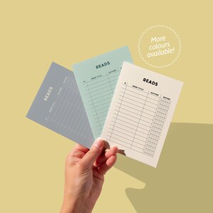 May include: Three different colored reading log trackers with the word "READS" at the top. The trackers are gray, light blue, and cream. Each tracker has columns for book title, author, and rating.