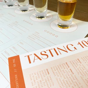 May include: A white tasting mat with four small glasses filled with clear liquid. The mat has a brown background with white text that reads "TASTING 10" and includes descriptions of different types of tequila: "A&ntilde;ejo", "Extra A&ntilde;ejo", and "Joven".