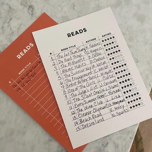 May include: A white sheet of paper with a handwritten list of book titles, authors, and ratings. The list is titled "Reads" and includes titles like "The Let Them Theory", "Do Hard Things", and "The Alchemist".