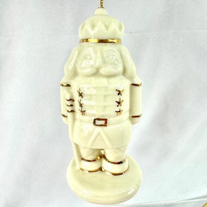 以下が含まれることがあります： 金色のアクセントが付いた白い磁器製のナッツクラッカーのオーナメント。ナッツクラッカーは帽子と金色のトリムのついたユニフォームを着ています。オーナメントは金色の紐で吊り下げられています。