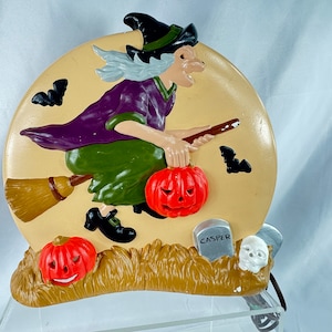 May include: A ceramic Halloween decoration of a witch flying on a broomstick with a pumpkin in her hand. The witch is wearing a purple dress and a green cape. The background is a tan moon with bats flying around it. The decoration has a tombstone that says "Casper" and a skull.