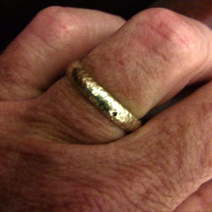 May include: A textured gold ring. The ring has a hammered or bark-like texture. The ring is on a finger, with the skin tone appearing light. The ring is a simple band.