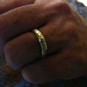 May include: Close-up of a textured gold ring on a hand. The ring has a hammered or organic design, and the metal has a slightly aged appearance. The ring is the primary focus, with the hand and a watch in the background.