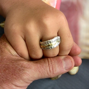 May include: A wide band ring featuring a textured, silver-colored exterior with a smooth, gold-colored center stripe. The ring has a rugged, hammered appearance across the silver sections, contrasting with the polished finish of the central band.