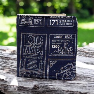 May include: A black fabric wallet with a map of Middle Earth from the Lord of the Rings. The wallet has a gold and white design with the text "LOTR MAP!" and "Facts about Middle Earth".