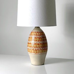 May include: A ceramic table lamp with a white drum shade. The lamp base is a cream color with a geometric pattern of orange circles.