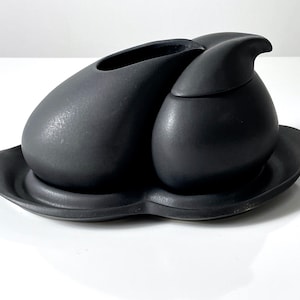 May include: A black ceramic sugar and creamer set with a unique, organic shape. The creamer has a lid with a spout. The set sits on a matching black ceramic tray.