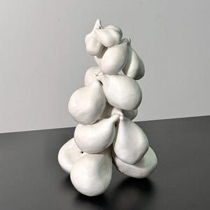 May include: A white ceramic sculpture of a cluster of teardrop-shaped forms. The forms are stacked together, creating a vertical arrangement. The sculpture is set against a neutral background.