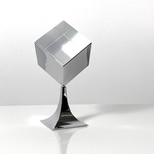 May include: A silver, geometric sculpture with a cube on top of a flared base.